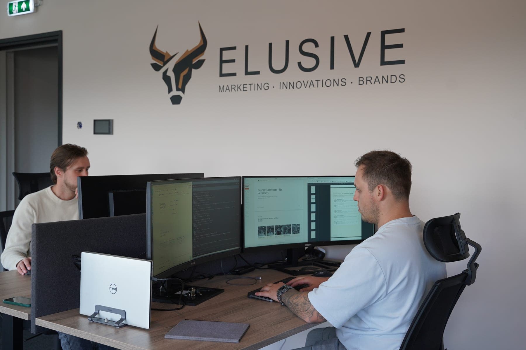 Twee teamleden werken aan hun bureau onder het Elusive-logo op de muur